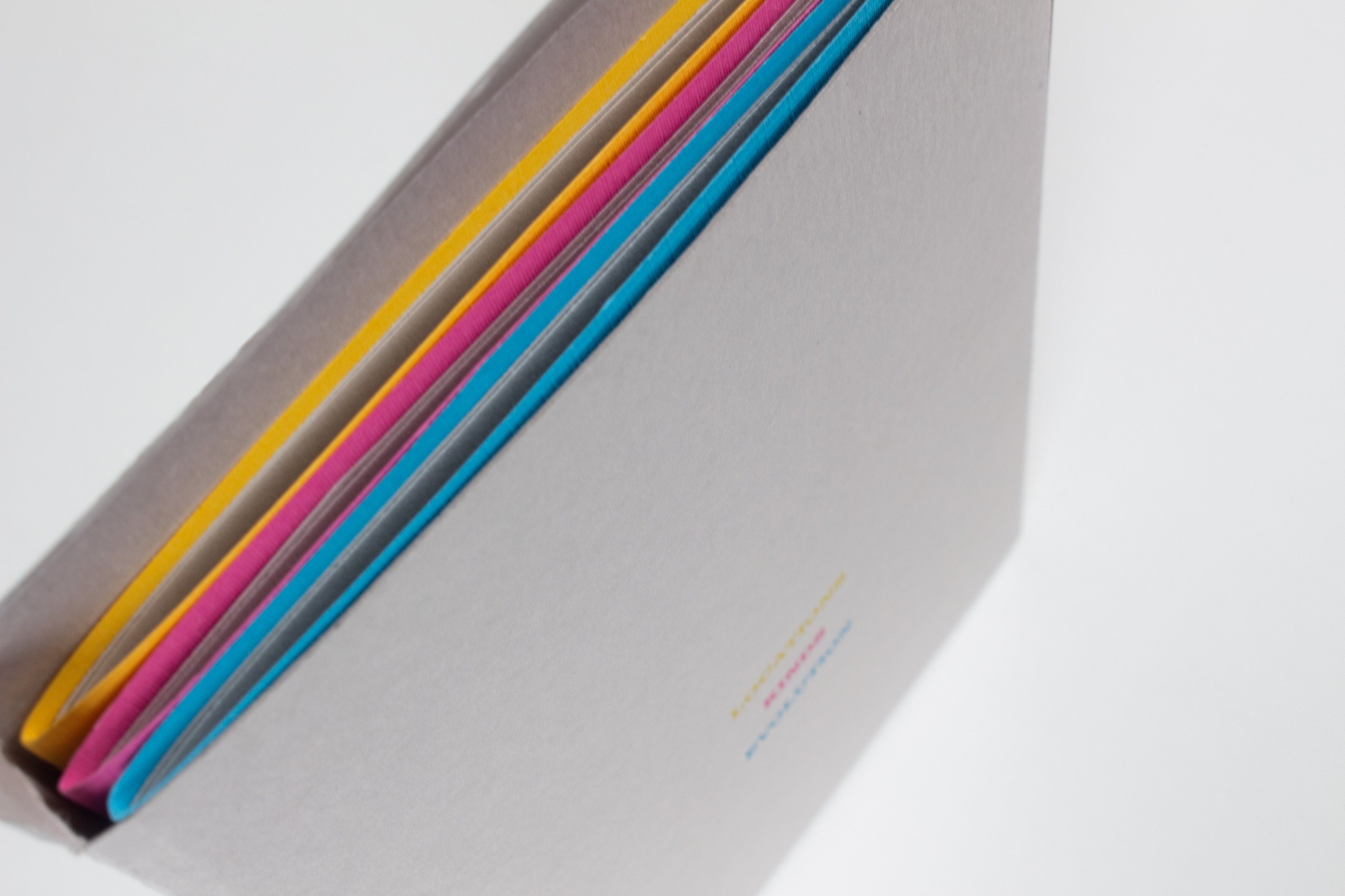 A raking close-up of the open end of the slipcase, the yellow, magenta and blue page edges of the three books stacked inside it