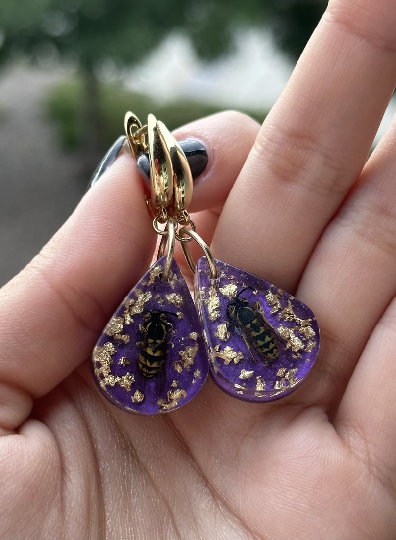 A pair of purple teardrop earrings, each with a bee
