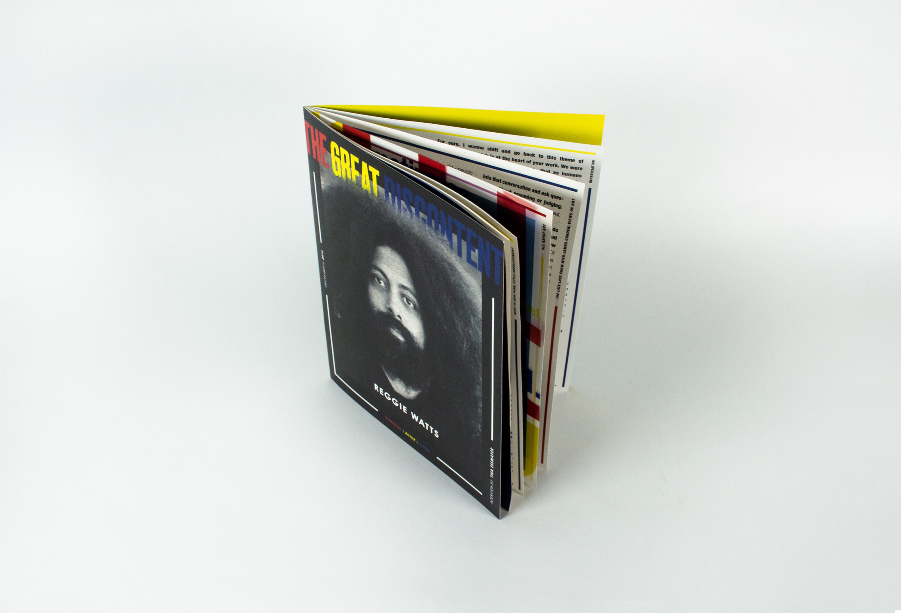 The closed magazine standing on a white surface, its fanned edges showing red, yellow and blue sections, a black-and-white portrait of Reggie Watts on the cover