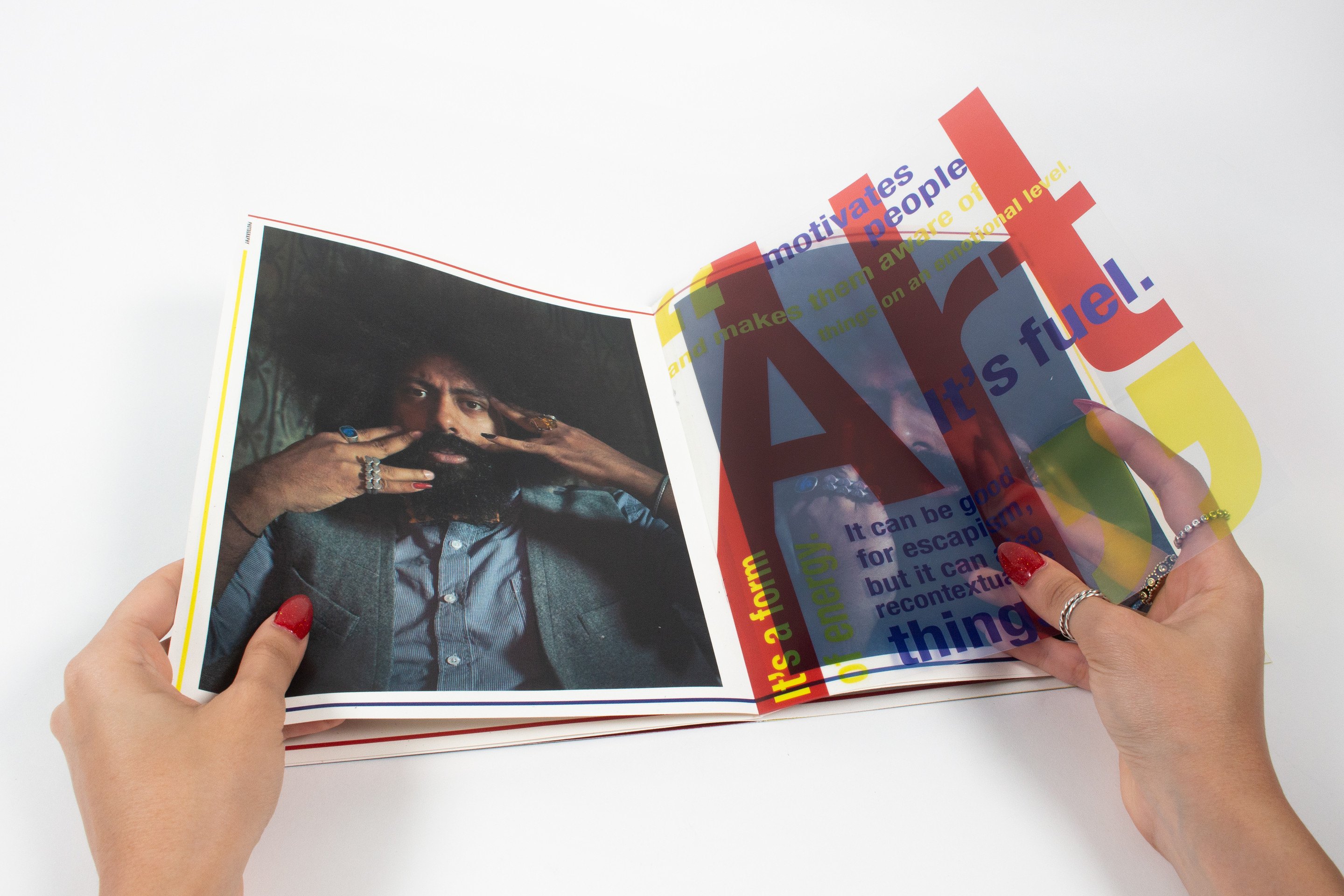 A hand turning the transparent sheet over the “Art” spread so the coloured type slides across the photograph beneath