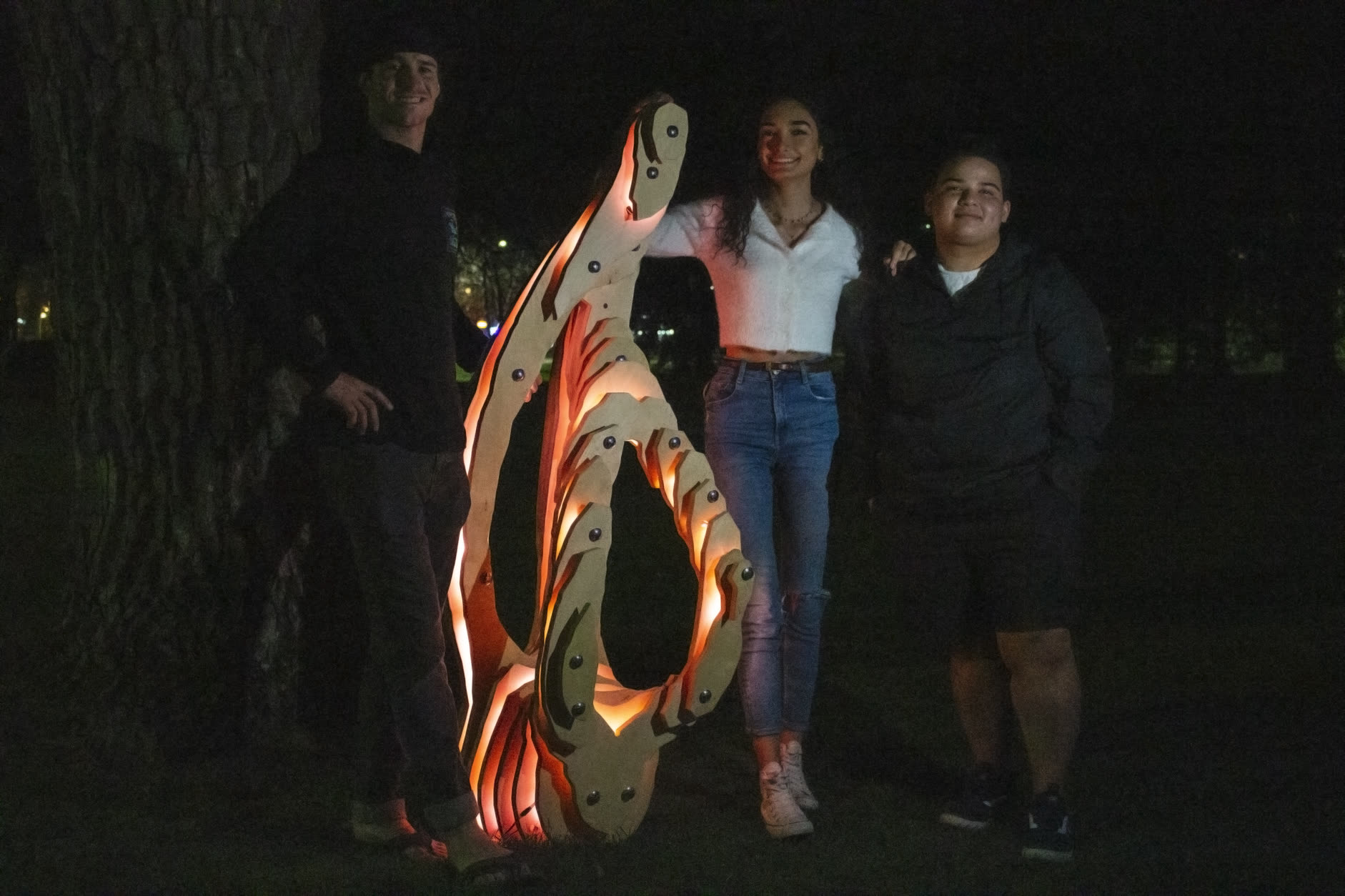 The three teammates standing either side of Zora on the lawn at night, the sculpture lit warm orange.